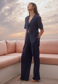 Ensemble deux pièces bleu marine composé d'une veste courte à manches courtes et décolleté en V profond, et d'un pantalon ample. Texture lisse, design minimaliste, sans attaches.