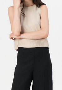 Beige, ärmelloses Oberteil mit rundem Ausschnitt und strukturierter Stoffqualität, kombiniert mit schwarzen Hosen mit weitem Bein. Das Oberteil hat eine lockere Passform und eine verkürzte Länge.