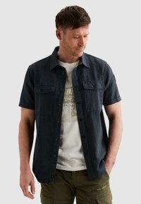 Homme portant une chemise à manches courtes bleu foncé boutonnée sur un t-shirt blanc à motif et un pantalon cargo vert olive, regardant vers le bas à sa gauche.