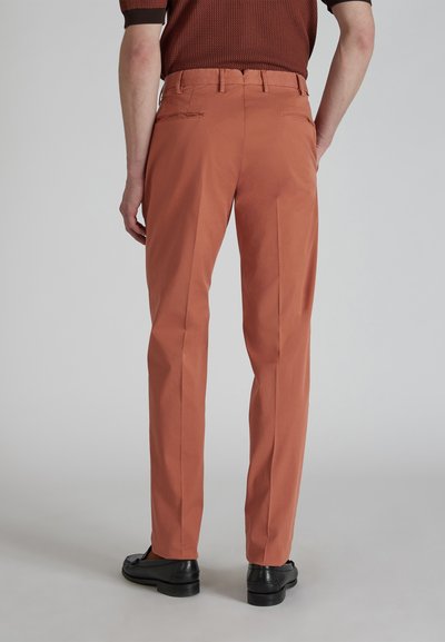 Pantaloni aderenti di colore ruggine realizzati in tessuto leggero. Presentano una parte frontale liscia, tasche laterali e una texture morbida. Le calzature sono nere.