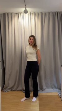 Joven mujer de pie descalza sobre suelo de madera, vistiendo camisa blanca y pantalones negros frente a cortinas beige cerradas bajo luz de techo.