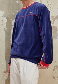 Modrá sametová mikina s červenými akcenty, s kulatým výstřihem a malým logem na hrudi. Měkká textura s uvolněným střihem.