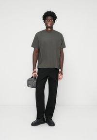 Dunkelgraues T-Shirt mit kurzen Ärmeln und lockerem Schnitt, kombiniert mit schwarzen Hosen und schwarzen Schuhen. Die Person trägt zudem eine kleine graue Tasche.