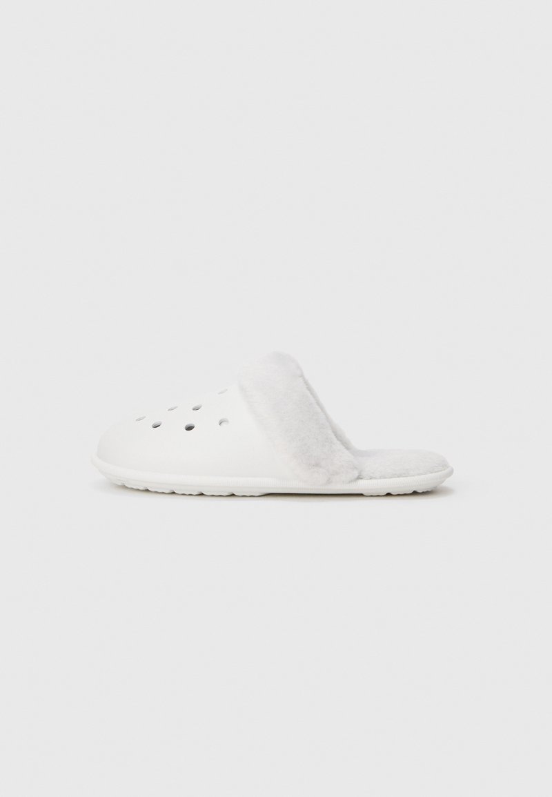 Vit slip-on klack med ventilationshål och grå plyschfoder på innersulan och kragen, visad från sidan mot en vit bakgrund.