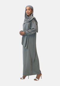 Lang, svajende grå kjole med lange ærmer og sammensnørede manchetter, parret med en matchende hijab. Båret med sølvfarvede høje sandaler. Glat tekstur.