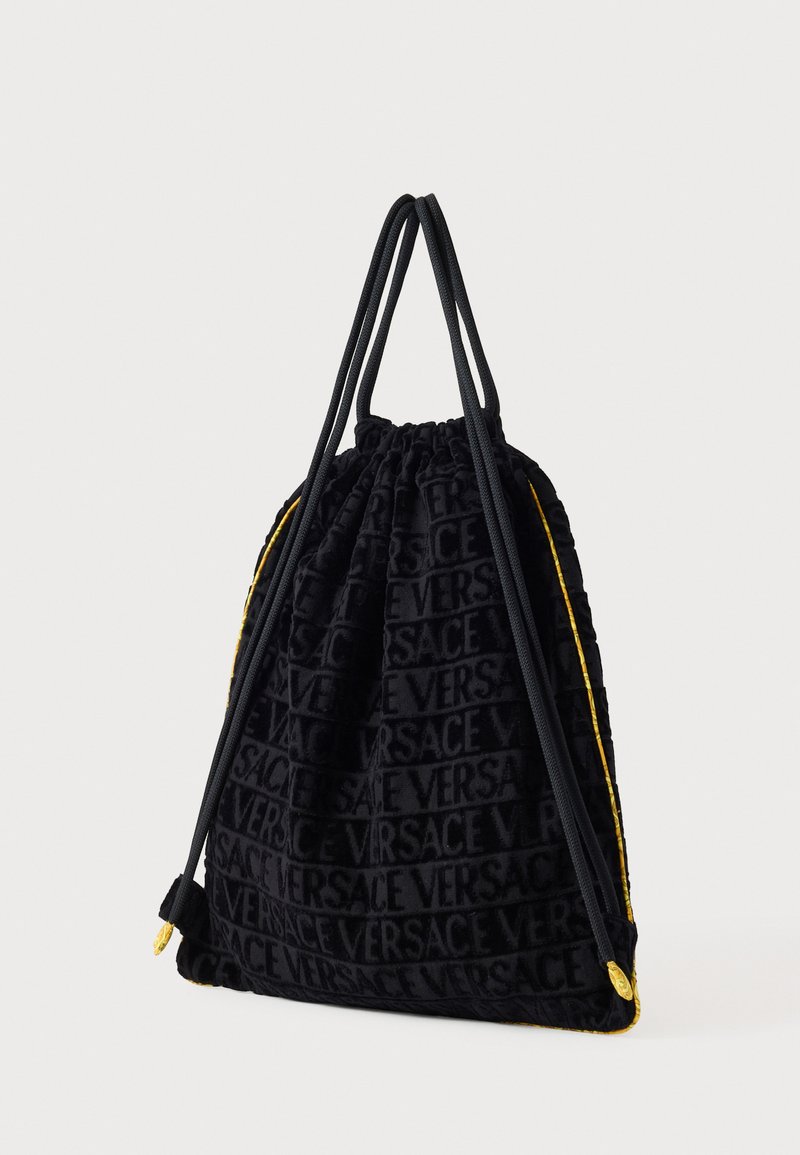 Sort fløjlsposse med snøre, prydet med prægede Versace-logo, gul kantliste og dobbelte sorte stropper, med en blød, struktureret finish.