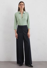 Light green button-up shirt with long sleeves, paired with high-waisted, wide-leg navy trousers and black footwear, set against a plain background.