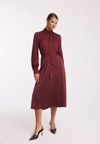 Burgundowa sukienka z długimi rękawami, wysokim kołnierzem i wiązaniem przy dekolcie. Wykonana z gładkiego materiału, sięgająca do połowy łydki, zestawiona z czubkowymi szpilkami.