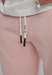 Gros plan sur un pantalon de survêtement rose avec des cordons blancs dotés d'embouts noirs et des inscriptions imprimées, associé à un vêtement zippé blanc.