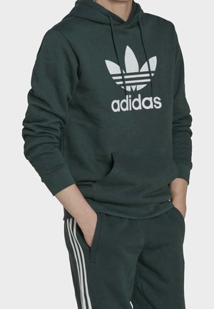 Person trägt dunkelgrünen Adidas-Hoodie mit großem weißem Trefoil-Logo und passende Hose mit weißen Seitenstreifen, Hände in den Taschen.