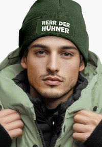 Green knit beanie with "HERR DER HÜHNERR" text and a lightly padded olive green jacket. Black inner lining visible at the collar.
