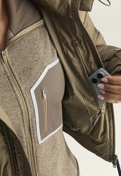 Mano sostiene un smartphone gris, colocándolo dentro de un bolsillo interior con cremallera de una chaqueta verde oliva sobre un forro polar beige con cremallera.