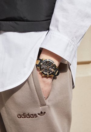 Personne portant un pantalon de jogging beige Adidas et une montre-bracelet noire et dorée, main glissée négligemment dans la poche du pantalon.