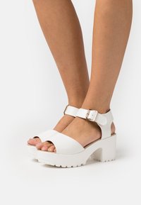 White chunky sandals with a smooth finish, featuring an ankle strap with a metal buckle and an open-toe design. Textured sole for grip.