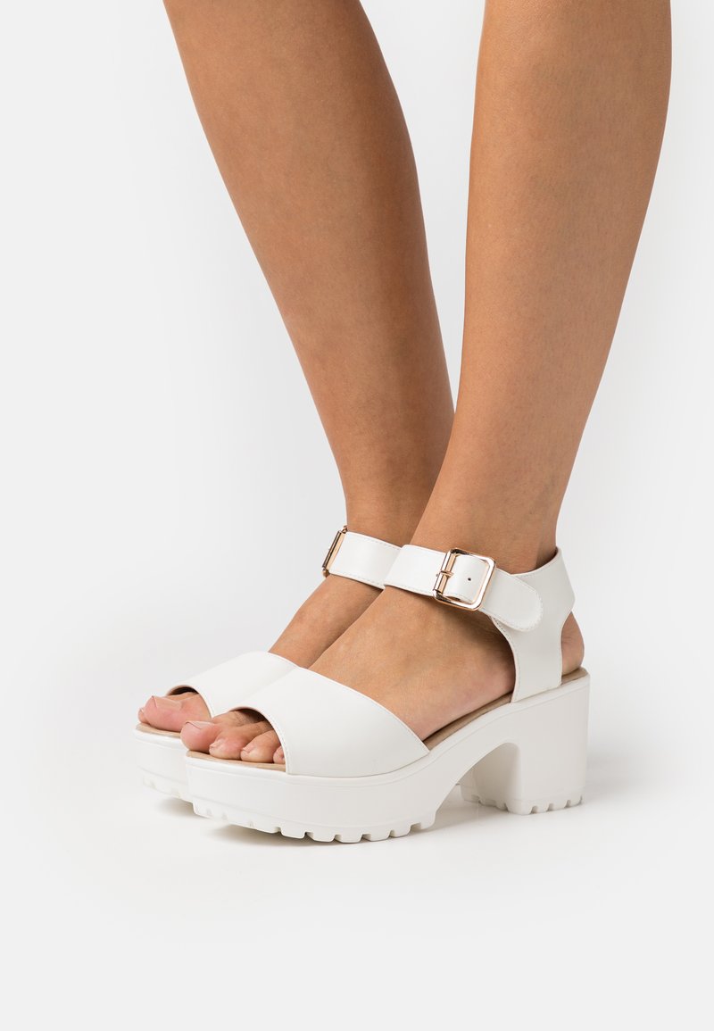 White chunky sandals with a smooth finish, featuring an ankle strap with a metal buckle and an open-toe design. Textured sole for grip.