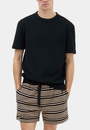 Mężczyzna w gładkim czarnym t-shircie z krótkim rękawem i czarno-beżowych, poziomo paskowanych szortach, z rękami w kieszeniach.