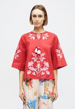 Γυναίκα που φοράει κόκκινο πουκάμισο με λευκή κεντητή ανθοδέσμη και κορδόνια με φούντες, συνδυασμένο με ανοιχτόχρωμο παντελόνι με πολύχρωμα σχέδια από θαλάσσια ζωή.