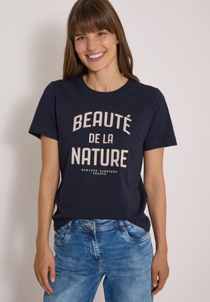 Femme souriante aux cheveux bruns et lisses porte un t-shirt bleu marine avec le texte "Beauté de la Nature" et un jean bleu sur un fond uni.