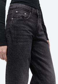 Μαύρο τζιν με φαρδιά μπατζάκια και ψηλή μέση, φορεμένο με μαύρο μακρυμάνικο τοπ, που δείχνει τις ραφές μπροστά και στο πλάι καθώς και μεταλλικά τρουκς στις τσέπες.