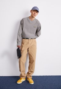 Sudadera gris con logo, pantalones de utilidad beige y zapatillas amarillas con rayas blancas. La persona sostiene un objeto acolchado oscuro.