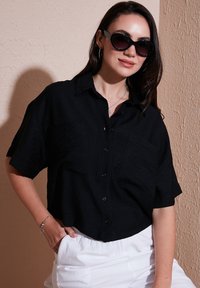 Camicetta nera a maniche corte con colletto e due tasche sul petto, realizzata in tessuto leggero, abbinata a pantaloni bianchi larghi.