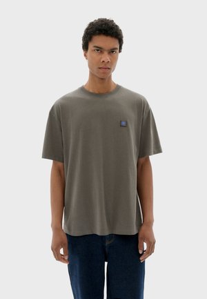 Olive green cotton t-shirt with a relaxed fit, short sleeves, and a small blue logo patch on the chest. Smooth texture, simple design.