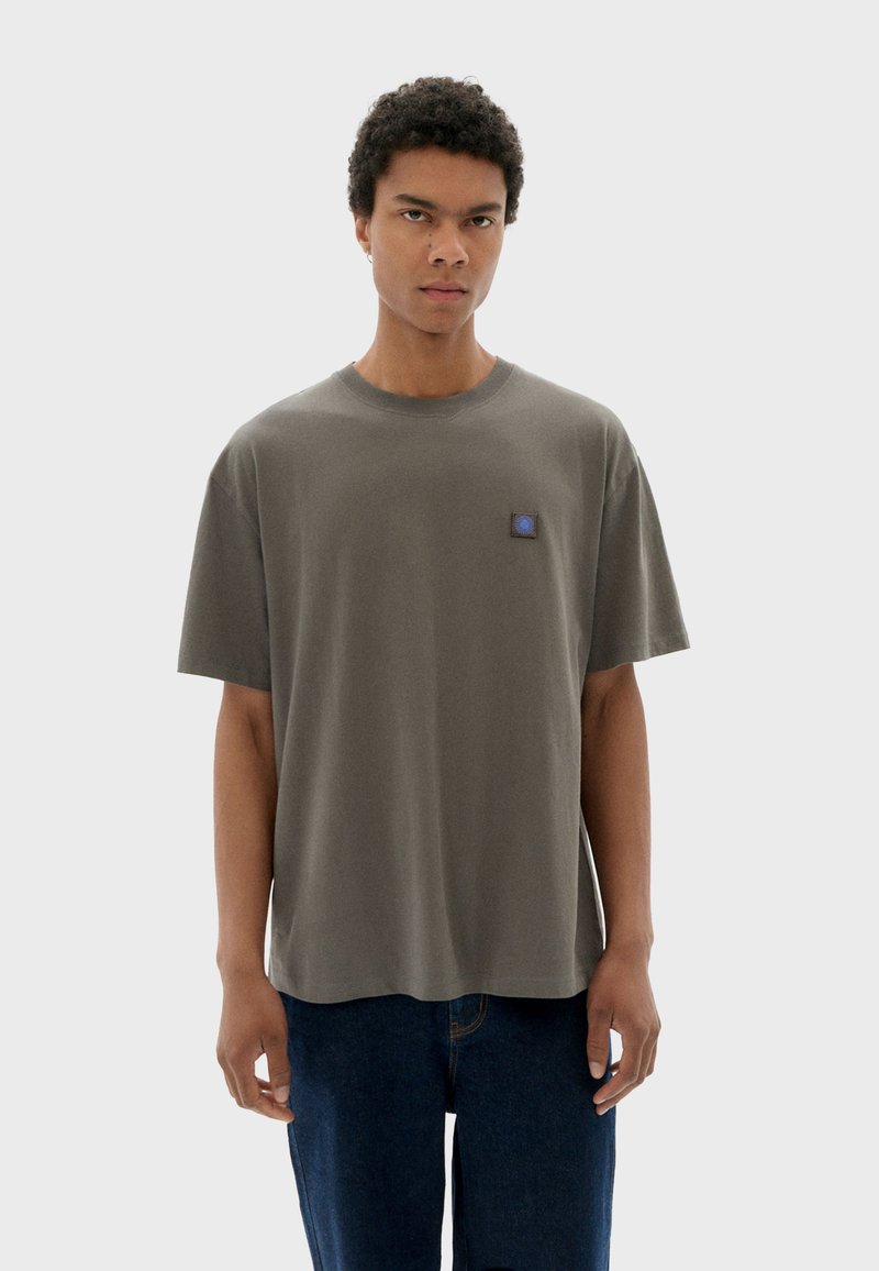 Olivgrünes Baumwoll-T-Shirt mit lockerer Passform, halblangen Ärmeln und einem kleinen blauen Logo-Patch auf der Brust. Glatte Textur, einfaches Design.