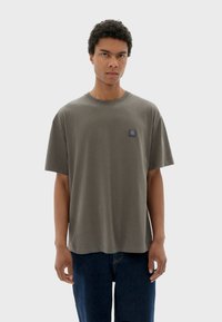 T-shirt à manches courtes olive en coton doux, avec un petit patch logo bleu sur la poitrine et une coupe décontractée.
