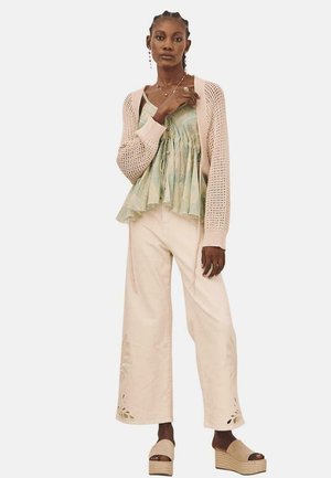 Cardigan de punto rosa claro sobre una blusa verde con estampado y lazo, combinado con pantalones de pierna ancha crema con recortes florales y sandalias de plataforma beige.