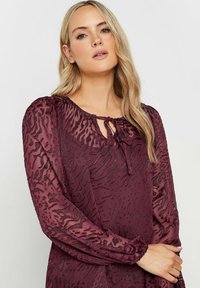 Blusa borgogna con stampa animalier testurizzata, maniche lunghe e scollatura a chiave. Realizzata in tessuto leggero e trasparente con polsini arricciati.