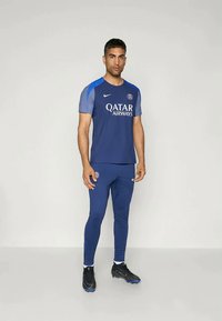 Muž stojí oblečený v modrom tréningovom obleku Paris Saint-Germain s logom Qatar Airways a čiernymi kopačkami na jednoliatom pozadí.