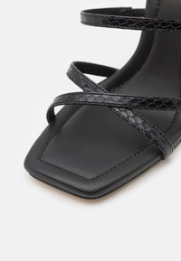 Sandale noire à bout carré avec de fines brides texturées croisées sur la semelle intérieure sur fond blanc.