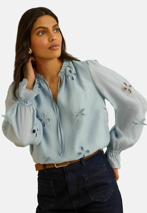 Woman wearing light blue blouse with floral cutouts and bows, dark pants, and brown belt, posing with hand near hair and looking up.