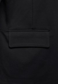 Poche de veste noire avec fermeture à rabat horizontale, bords cousus et lignes de couture subtiles sur une surface de tissu lisse.