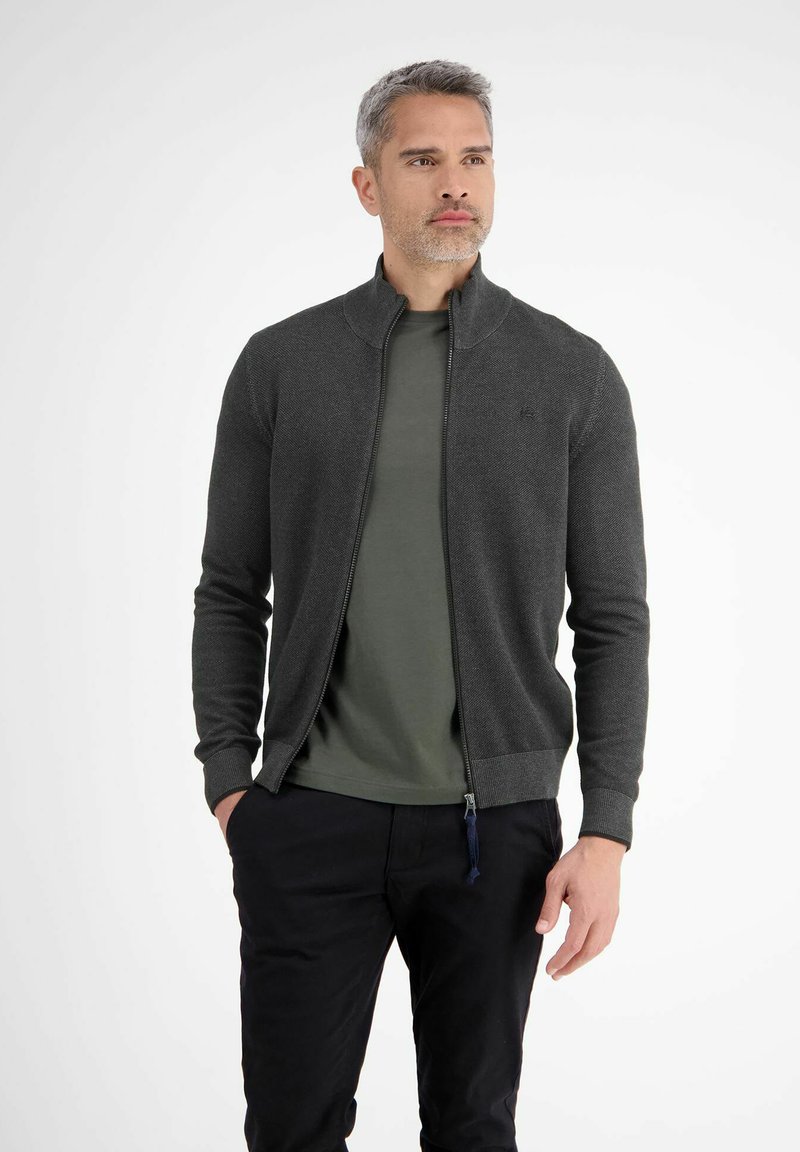 Mann mit grauem Haar, der einen dunkelgrauen Reißverschlusscardigan über einem grünen Shirt und schwarzen Hosen trägt, steht mit den Händen in den Taschen.