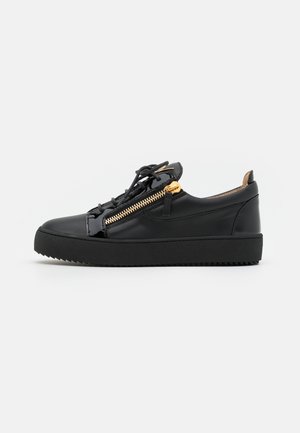 Sapatilhas de couro preto com uma sola de borracha texturizada, apresentando um detalhe em zíper dourado e fecho de atacadores. Acabamento liso e design de baixo perfil.