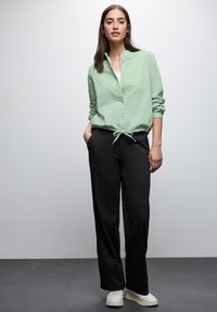 Camicia verde chiaro a maniche lunghe con bottoni e vita regolabile, abbinata a pantaloni neri a gamba larga e sneakers bianche. Design semplice.