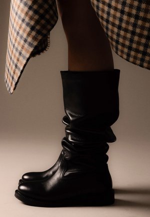 Bottes en cuir noir souples mi-mollet portées avec une jupe à carreaux beige et marine sur un fond neutre uni.