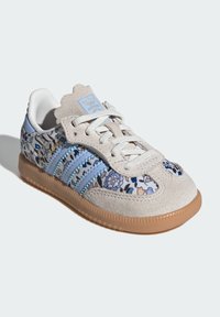 Basket Adidas Samba avec suède beige, tissu floral bleu, rayures bleu clair, lacets blancs et semelle en caoutchouc, positionnée en biais à gauche sur un fond blanc.