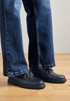Chaussures en cuir bleu marine avec une semelle épaisse et un détail en chaîne sur le dessus, assorties à un jean en denim bleu foncé à coupe large.