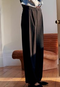 Pantaloni scuri, a fantasia, con una vestibilità ampia e pieghe nette. Abbinati a scarpe basse nere, in piedi su un pavimento di legno vicino a un divano curvo rosa.
