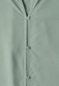 Chemise vert clair avec fermeture à boutons verticale et col en V montrant quatre boutons, dont un avec un texte visible.
