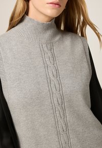 Suéter gris de punto con un alto cuello acanalado, que presenta detalles de punto cableado en el centro y mangas de tela negra. Textura suave.