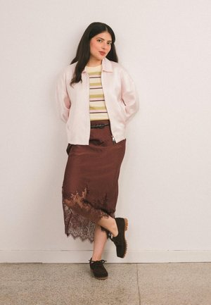 Vrouw met lang donker haar staat tegen een muur, gekleed in een roze satijnen jas, een gestreept topje, een midi-rok met bruine kantaccenten en zwarte schoenen.