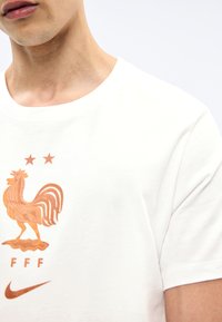 Κοντινό πλάνο ενός ατόμου που φοράει ένα λευκό πουκάμισο με έμβλημα χάλκινου κόκορα, δύο αστέρια, τα γράμματα "FFF" και το λογότυπο της Nike κάτω από το σχέδιο.