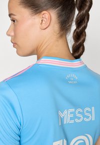 Svijetloplava sportska majica s okruglim izrezom, ružičasto-bijelim prugama na ovratniku i ispisom "MESSI" u bijeloj boji.