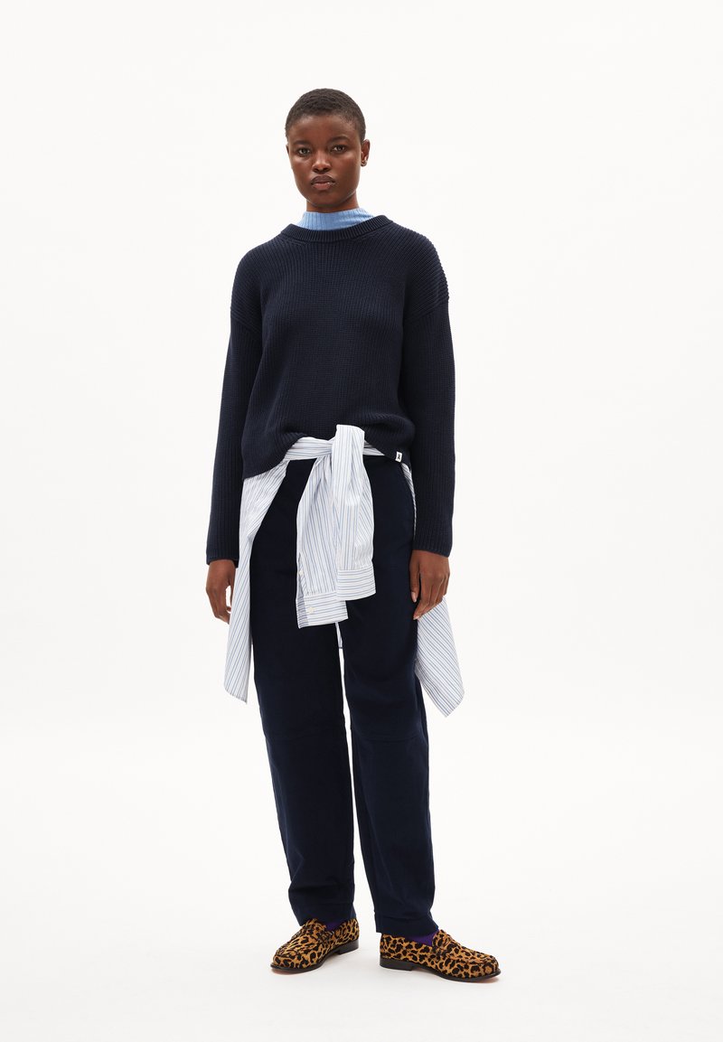 Marineblauer Strickpullover, hellblaues Hemd mit Kragen, marineblaue Hose und Hausschuhe mit Leopardenmuster; das Modell posiert mit einem gebundenen gestreiften Hemd.
