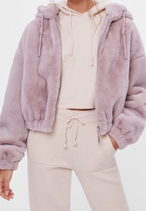 Mujer con chaqueta borrosa de cremallera de color rosa pálido sobre sudadera con capucha crema y pantalones crema a juego con cordón, con la mano en el bolsillo.