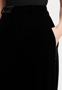 Pantalons taille haute en velours noir avec poches latérales, présentant une texture lisse et une coupe ajustée. Détails de couture le long des côtés.
