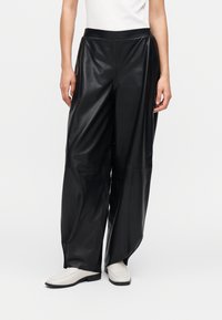 Calças de ganga largas pretas em couro falso, com cintura alta, textura suave e costuras subtis. Combinadas com sapatos deslizantes de cor clara.